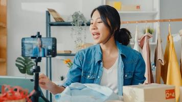 jonge aziatische dame modeontwerper met behulp van mobiele telefoon die inkooporder ontvangt en kleding laat zien die video livestreaming online opneemt in de winkel. eigenaar van een klein bedrijf, online marktleveringsconcept. foto