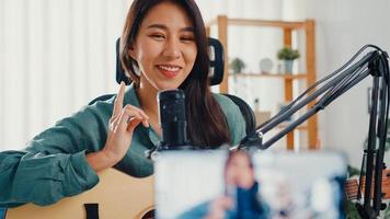 tiener azië meisje influencer gitaar spelen muziek gebruik microfoon record met smartphone voor online publiek luister thuis. vrouwelijke podcaster maakt audiopodcast vanuit haar thuisstudio, blijf thuis concept.