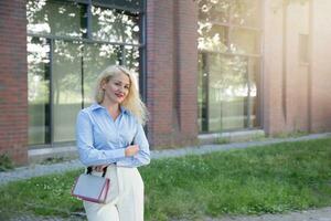 een zakenvrouw in een blauw overhemd en wit broek is staand buiten de kantoor. een warm zonnig dag. een jong vrouw staat in de buurt een rood gebouw. foto