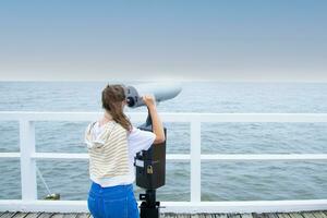 een jong vrouw staat Aan de pier, op zoek door munt verrekijker. achterzijde visie. Baltisch zee, verkoudheid weer foto