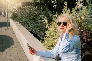 een blond vrouw is zittend Aan een bank Aan de straat en gebruik makend van een telefoon. een mooi vrouw in een blauw overhemd en wit broek, zonnebril. zomer dag foto