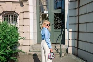 een jong vrouw gekleed in een blauw overhemd en wit broek is wandelen door de stad naar de kantoor. blond haar, zonnebril. warm zomer het weer. visie van de terug foto