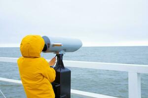 de jongen is staand Aan de pier, op zoek door kijker Bij de Baltisch zee. verkoudheid het weer. grijs lucht foto