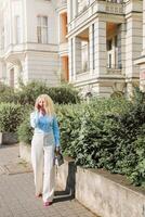 een blond vrouw wandelingen naar beneden de straat en praat Aan de telefoon. een mooi vrouw in een blauw overhemd en wit broek, zonnebril. zomer dag foto