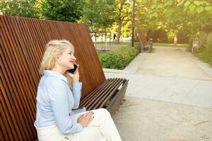 een mooi hoor jong vrouw is zittend Aan een bank in de park, pratend Aan de telefoon, video communicatie. zomer dag. kant visie foto