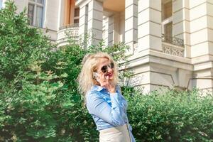 een blond vrouw wandelingen naar beneden een stad straat en praat Aan de telefoon. een mooi vrouw in een blauw overhemd en wit broek, zonnebril. zomer dag foto