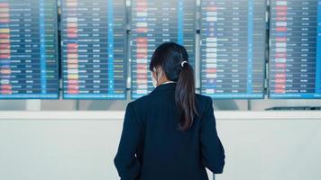 aziatisch zakenmeisje draagt een gezichtsmasker met een kofferstandaard voor het bord, kijk naar informatie die haar vlucht controleert op de internationale luchthaven. zakelijke forens covid pandemie, zakelijk reisconcept. foto