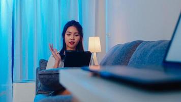 Aziatische zakenvrouw die tablet gebruikt, praat met collega's over het plan in een videogesprek terwijl ze 's nachts vanuit huis in de woonkamer werkt. zelfisolatie, sociale afstand, quarantaine voor coronaviruspreventie.