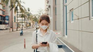 succesvolle jonge Aziatische zakenvrouw in mode-kantoorkleding met een medisch gezichtsmasker met behulp van een smartphone terwijl ze 's ochtends alleen buiten in de stedelijke moderne stad loopt. bedrijf onderweg concept. foto