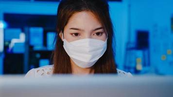 gelukkige zakenvrouw in azië die een medisch gezichtsmasker draagt voor sociale afstand in een nieuwe normale situatie voor viruspreventie tijdens het gebruik van een laptop op het werk in de kantoornacht. leven en werken na het coronavirus. foto