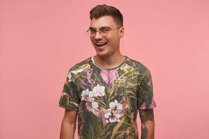 jong mooi vent op zoek Bij camera met breed glimlach, wezen positief en Vrolijk, vervelend gebloeid t-shirt en bril, staand over- roze achtergrond foto