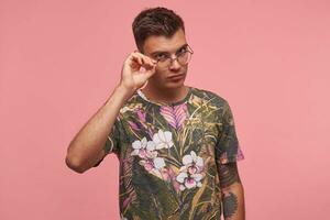 detailopname van echt mooi vent poseren over- roze achtergrond, op zoek Bij camera met zwaar zicht, gaan naar naar nemen zijn eyewear uit, vervelend t-shirt met bloemen afdrukken foto