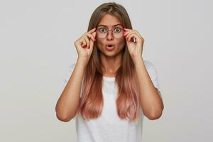 verbijsterd jong bruine ogen lang haren blond dame houden haar eyewear met verheven handen terwijl op zoek verbaasd naar camera, gekleed in wit eenvoudig t-shirt terwijl poseren over- wit achtergrond foto