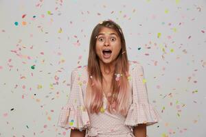 binnen- schot van jong lief verrast lang haren vrouw op zoek Bij camera verbaasd terwijl staand over- wit achtergrond en kleurrijk confetti, gekleed in feestelijk kleren foto