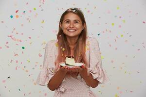 portret van vrolijk jong lief vrouw met licht bruin lang haar- poseren in veelkleurig confetti met stuk van taart in verheven handen, op zoek omhoog en glimlachen aangenaam foto