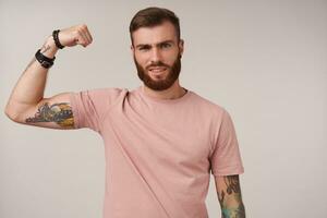 knap jong brunette Mens met baard en tatoeages verhogen hand- en tonen zijn stroom, op zoek Bij camera met verheugd gezicht en loensen, poseren over- wit achtergrond in gewoontjes kleren foto