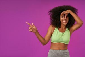 portret van jong gekruld donker huid vrouw met gewoontjes kapsel staand over- Purper achtergrond, op zoek Bij camera en sluitend neus- met hand, vermijden slecht geur en tonen terzijde met inhoudsopgave vinger foto