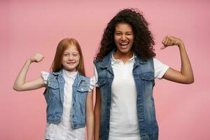 twee vrolijk mooi jong Dames staand tegen roze achtergrond met verheven handen en demonstreren gelukkig hun sterk biceps, vervelend jeans vesten en wit overhemden foto