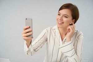 portret van aangenaam op zoek jong bruin haren vrouw met kort modieus kapsel glimlachen positief terwijl hebben video babbelen met mobiel telefoon en koptelefoon, geïsoleerd over- wit achtergrond foto