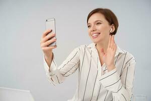 vrolijk jong kort haren brunette vrouw met natuurlijk bedenken verhogen hand- in Hallo gebaar en glimlachen aangenaam terwijl maken video telefoontje met haar smartphone, geïsoleerd over- wit achtergrond foto