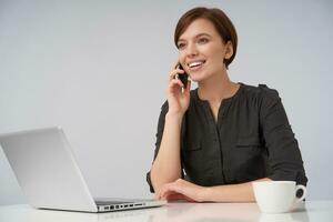 vrolijk jong bruine ogen lief brunette vrouw met kort modieus kapsel glimlachen aangenaam terwijl maken telefoongesprek, zittend Bij tafel over- wit achtergrond met laptop en kop van koffie foto