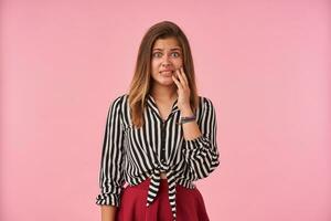 verbijsterd jong mooi brunette vrouw gekleed in feestelijk kleren aanraken haar gezicht met verheven hand- en afronding haar ogen terwijl op zoek Bij camera met steenbolk, geïsoleerd over- roze achtergrond foto