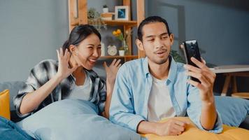 gelukkig jong aziatisch stel man en vrouw zitten op de bank en gebruiken smartphone facetime videogesprek met vrienden en familie in de woonkamer thuis. blijf thuis quarantaine, sociale afstand, jong getrouwd concept. foto