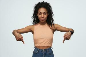 zelfverzekerd aantrekkelijk Afrikaanse Amerikaans jong vrouw staand en richten naar beneden door twee vingers Bij beide handen geïsoleerd over- wit achtergrond foto