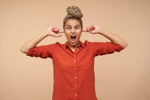 geërgerd jong groene ogen bruin haren vrouw schreeuwen stressvol terwijl inhammen haar oren met inhoudsopgave vingers, staand over- beige achtergrond in gewoontjes slijtage foto