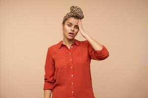 van streek jong mooi bruin haren vrouw met hoofdband loensen haar ogen en houden verheven palm Aan voorhoofd terwijl staand over- beige achtergrond in gewoontjes kleren foto