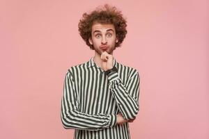 portret van jong groene ogen gekruld gebaard Mens verhogen verbaasd wenkbrauwen en achtervolgen zijn lippen terwijl op zoek Bij camera, houden verheven hand- Aan zijn kin terwijl staand over- roze achtergrond foto