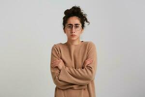 echt dame, fronsen vrouw met donker gekruld haar- broodje. vervelend beige jumper en bril. emotie concept. houdt armen gekruiste Aan een borst. aan het kijken Bij de camera geïsoleerd over- wit achtergrond foto