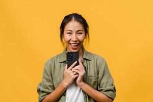 verraste jonge azië dame met behulp van mobiele telefoon met positieve uitdrukking, breed glimlachen, gekleed in casual kleding en camera kijken op gele achtergrond. gelukkige schattige blije vrouw verheugt zich over succes.