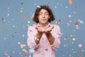 portret van Mens gelukkig staand in de omgeving van vallend naar beneden confetti, over- blauw achtergrond. jong Mens blazen confetti uit zijn handen. foto