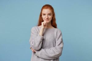 mooi speels roodharig meisje staand Holding hand- in de buurt de kin looks zelfverzekerd, glimlachend een beetje denken van iets prettig, sympathiek naar gesprekspartner, vervelend een warm trui foto