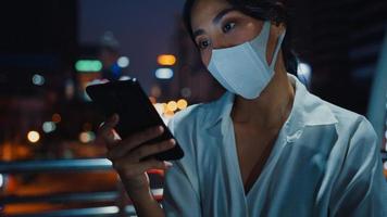 jonge azië zakenvrouw in mode kleding met gezichtsmasker met behulp van smartphone sms-bericht te typen terwijl je 's nachts buiten in de stedelijke stad staat. sociale afstand om verspreiding van het covid-19-concept te voorkomen.