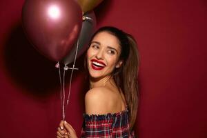 blij jong mooi donker haren vrouw met avond bedenken houden lucht ballonnen in verheven hand- en glimlachen op grote schaal, vervelend elegant top terwijl poseren over- bordeaux achtergrond foto