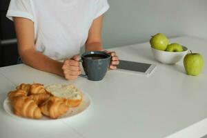 dichtbij omhoog van wit keuken tafel, visie van een kant, vrouw handen houdt een grijs kop met sommige vloeistof, croissants Aan een bord, appels in een schaal, telefoon met zwart scherm zijn Daar Aan een tafel foto