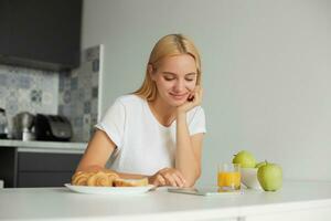 vrouw zit Bij de keuken tafel in de ochtend, lachend, looks Bij tablet zwart scherm, lezing bericht, Aan de tafel - een glas van sap, appels, broodjes, gekleed in een huiselijk t-shirt, keuken achtergrond foto