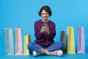 positief jong aantrekkelijk gekruld brunette vrouw Holding mobiel telefoon in verheven handen en op zoek vrolijk Aan scherm, zittend over- blauw achtergrond met papier Tassen foto