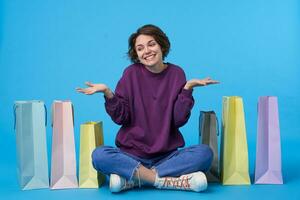 positief jong lief donker haren vrouw met gewoontjes kapsel schouderophalend met verheven palmen en glimlachen vrolijk, gekleed in Purper trui, jeans en sportschoenen terwijl zittend over- blauw achtergrond foto