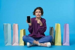 verbijsterd jong mooi kort haren gekruld vrouw Holding mobiel telefoon in verheven hand- en tonen verward Aan scherm, poseren over- blauw achtergrond met gekruiste poten foto