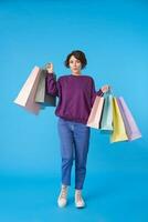 binnen- foto van jong mooi kort haren gekruld vrouw met papier Tassen in verheven handen terwijl poseren over- blauw achtergrond in Purper sweater en jeans