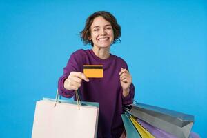 vrolijk jong lief brunette vrouw met kort kapsel wezen gelukkig terwijl hebben boodschappen doen en houden credit kaart in verheven hand, geïsoleerd over- blauw achtergrond foto