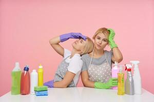studio foto van jong blond vrouwtjes gekleed in eenvoudig t-shirts en schorten zittend over- roze achtergrond met huishouden Chemicaliën, schoonmaak groot huis en wezen heel moe