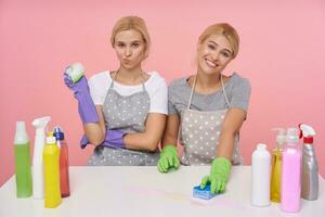 binnen- schot van jong withoofdig zussen in uniform en rubber handschoenen houden sponzen in hun handen terwijl schoonmaak tafel, zittend over- roze achtergrond foto