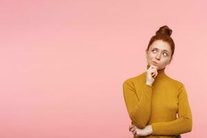 jong dame, zich afvragen vrouw met gember haar, sproeten en broodje. vervelend goud coltrui trui en aanraken haar kin in gedachten. aan het kijken naar de links Bij kopiëren ruimte over- pastel roze achtergrond foto