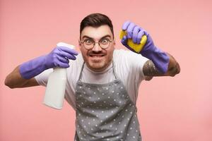 studio schot van jong blauwe ogen brunette mannetje in bril grimassen zijn gezicht terwijl op zoek Bij camera en houden huishouden Chemicaliën in zijn handen, geïsoleerd over- roze achtergrond foto