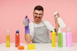 vrolijk jong knap kort haren brunette vent in werken kleren en rubber handschoenen hebben pret met verstuiven flessen terwijl maken breken met voorjaar schoonmaak, poseren over- roze achtergrond foto