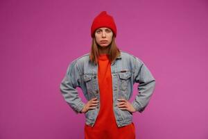 portret van jong vrouw met brunette lang haar. vervelend jeans jasje, rood trui en hoed. houdt handen Aan een taille, frons. aan het kijken verdacht Bij de camera geïsoleerd over- Purper achtergrond foto